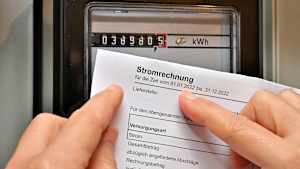 Eine Frau hält die Stromrechnung in der Hand und vergleicht diese mit dem Zählerstand an dem Stromzähler im Haus
