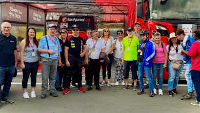 Eine Gruppe von Menschen steht vor einem roten Lastwagen, umgeben von Rennsport-Equipment. Der Himmel ist blau mit ein paar Wolken.