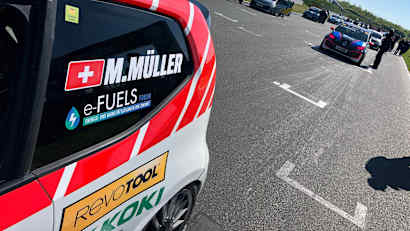 Eine Rennstrecke mit mehreren Autos unter blauem Himmel ist zu sehen. Links in Vordergrund steht ein Auto mit einem eFuels Forum Sticker.