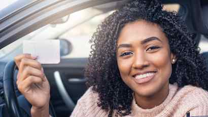 Eine lächelnde Frau sitzt im Auto und hält ihren Führerschein in die Kamera.
