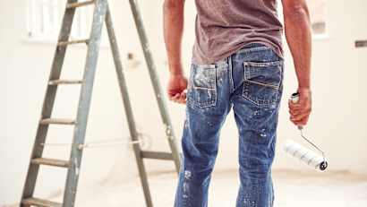 Close Up eines Mannes von hinten, welcher eine farbbemalte Hose trägt und eine Farbrolle in der Hand hält