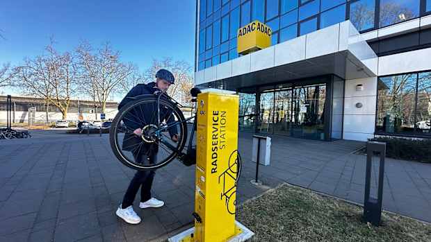 Ein Mann hängt ein Fahrrad an eine gelbe Säule mit der Aufschrift Radservicestation. Im Hintergrund ist ein verglastes Hochhaus mit ADAC Logo zu sehen.