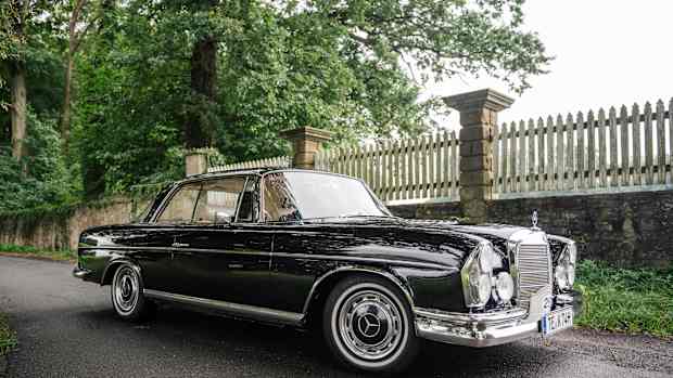 Ein schwarzer Oldtimer von Mercedes steht auf einer Straße. Im Hintergrund sieht man einen Zaun und einen Baum.
