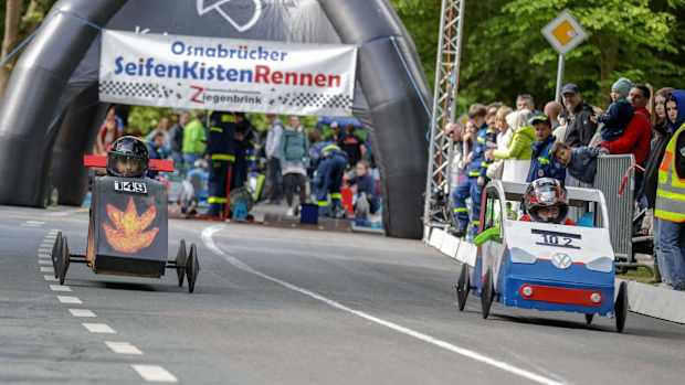 Auf einer Rennstrecke fahren zwei Seifenkisten nebeneinander. Im Hintergrund ist das Publikum zu sehen.