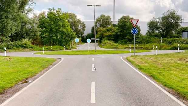 Eine leere Straße führt auf einen Kreisel hin, an dem Verkehrs- und Straßenschilder stehen.