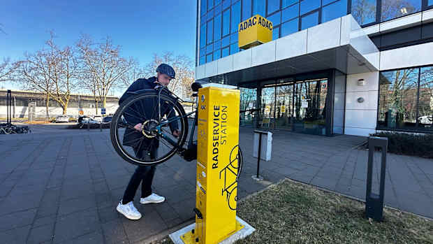 Ein Mann hängt ein Fahrrad an eine gelbe Säule mit der Aufschrift Radservicestation. Im Hintergrund ist ein verglastes Hochhaus mit ADAC Logo zu sehen.
