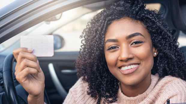 Eine lächelnde Frau sitzt im Auto und hält ihren Führerschein in die Kamera.