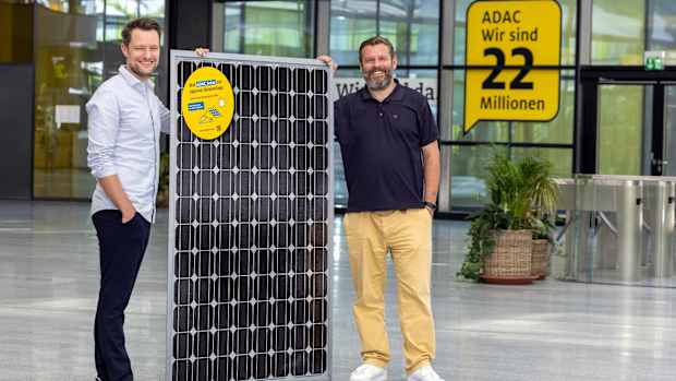 Zwei Männer halten ein Solarpanel aufrecht in dem Foyer eines Gebäudes und verdeutlichen so die Zusammenarbeit zwischen adac und otovo.