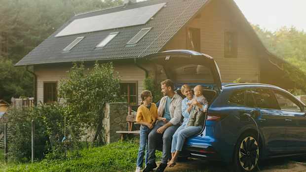 Eine Familie, die sich an einem sonnigen Tag vor ihrem E-Auto und Haus mit PV-Anlage aufhalten.s mit PV-Anlage sitzt.