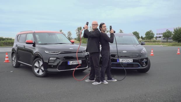 Dr. E und Thomas M. Held stehen Rücken an Rücken mit Ladekabeln für ihre E-Autos in der Hand und bereiten sich auf die Drift-Challenge vor.