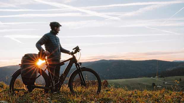 Ein Mann steht mit seinem E-Bike auf einem Hügel und schaut über eine Landschaft während die Sonne untergeht, ein E-Bike kaufen ist also eine gute Entscheidung.