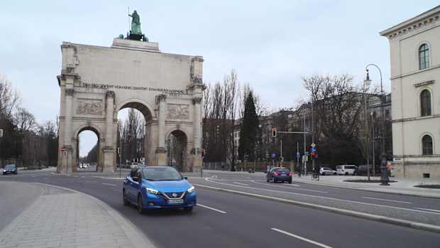 Ein blauer Nissan fährt vor dem Siegestor in München und zeigt wir man in der Stadt Strom sparen kann.