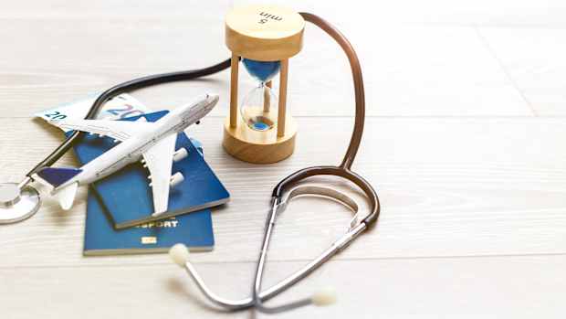 Stethoskop, Sanduhr, Flugzeugmodell, Reisepässe und Geld auf hellem Holzboden als Symbol für einen medizinischen Notfall auf Reisen.