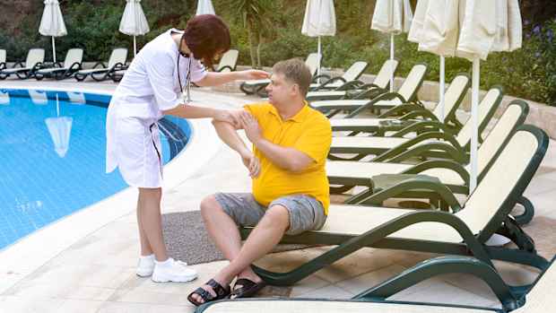 Eine Aerztin behandelt einen maennlichen Reisenden, der auf einer Liege am Pool eines Hotels sitzt. Im Hintergrund stehen weitere Liegen und Sonnenschirme.