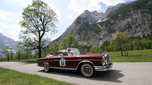 Zwei Männer fahren in einem roten Oldtimer eine Straße neben den Bergen entlang und müssen sich keine Gedanken über eine Oldtimerversicherung machen