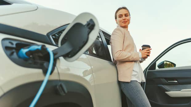 Eine Frau steht angelehnt an ein weißes E-Auto und sieht ihm beim laden zu, laut einer Umfrage wollen viele Menschen ein E-Auto kaufen