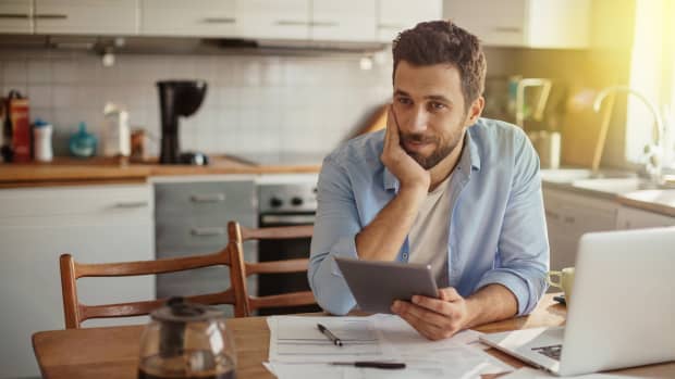 Ein Mann sitzt an seinem Esstisch mit einem Tablet in der Hand, vielen verteilten Dokumenten und einem Laptop und macht sich Gedanken über seine Rechtsschutzversicherung