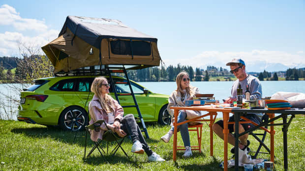 Eine Gruppe junger Leute sitzen an einem Campingtisch vor einem aufgebauten Dachzelt an einem See