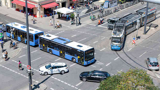 Verschiedene Verkehrsteilnehmer wie Autofahrer, Straßenbahn, Bus, Fußgänger und Radfahrer fahren über eine Kreuzung