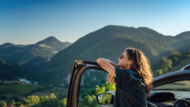 Eine Frau steht an ihrer Autotür gelehnt im Sommer mit Ausblick auf die Berge