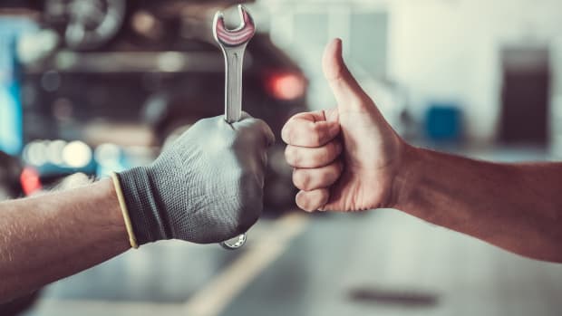 Zwei Männer zeigen einen Schraubenschlüssel und ein Daumen-Hoch Zeichen. Im Hintergrund eine Autowerkstatt