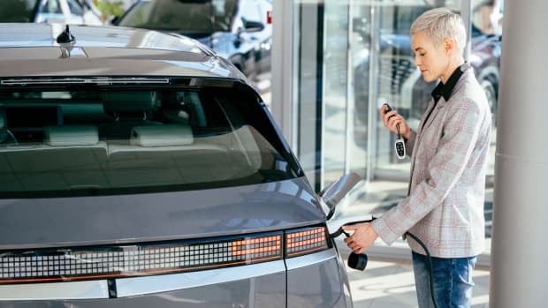 Frau steht in der Nähe von Elektroautos mit Fernbedienung in der Hand im Autohaus. Frau mit Ladekabel für Elektroauto, glückliches Warten auf Elektroauto zum Aufladen.