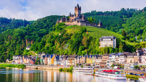 Eine Stadt an einem Fluss, mit Schiffen und über der Stadt liegt eine Burg auf einem Berg