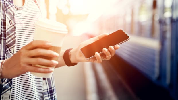 Eine Frau steht mit ihrem Smartphone und einen Coffee-to-go Becher vor einem Zug