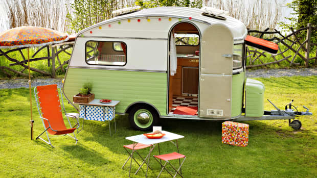 Grünweißer Retro Wohnwagen auf Wiese mit orangefarbenem Sonnenstuhl, Tisch, Hocker und Kühbox davor ..