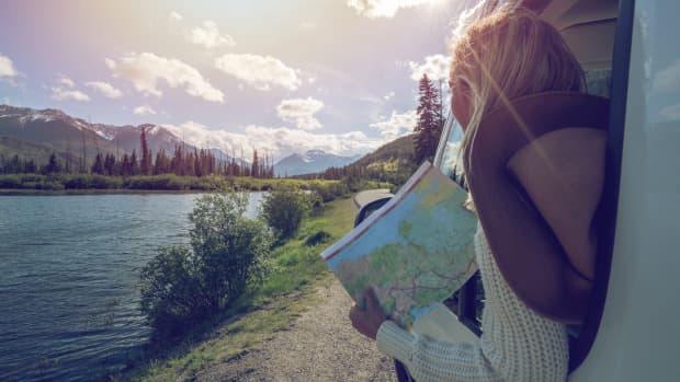 Eine Frau hat eine Landkarte in der Hand und lehnt sich aus einem Auto und schaut auf Berge und auf einen See.