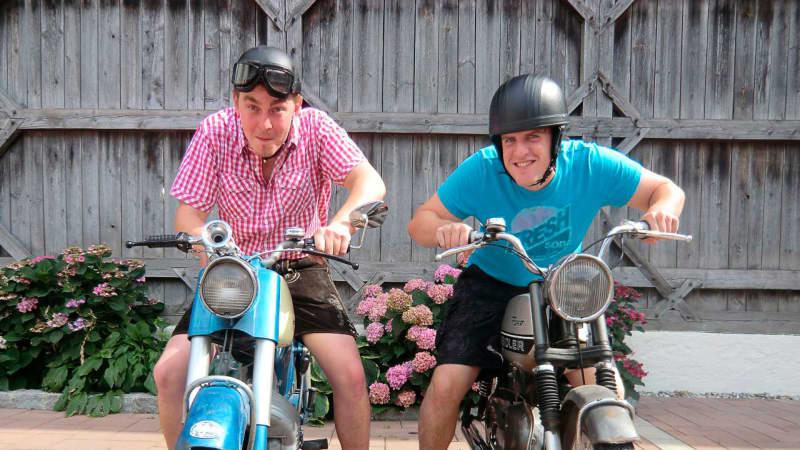 Zwei Männer auf alten Motorrädern mit alten Helmen blicken in die Kamera
