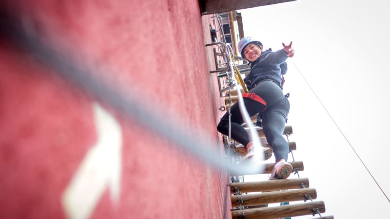 Eine Frau mit Helm und Sicherung klettert eine Hängeleiter an einem hohen Gebäude hinauf