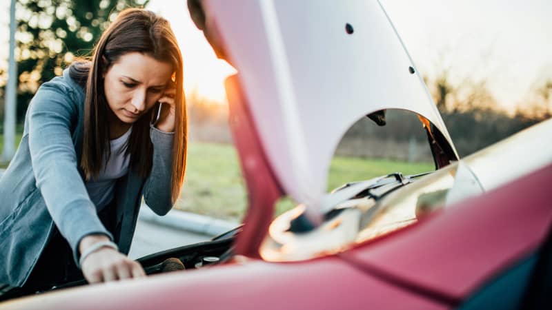 Junge Frau telefoniert beim Blick in die Motorhaube weil ihr Auto nicht anspringt