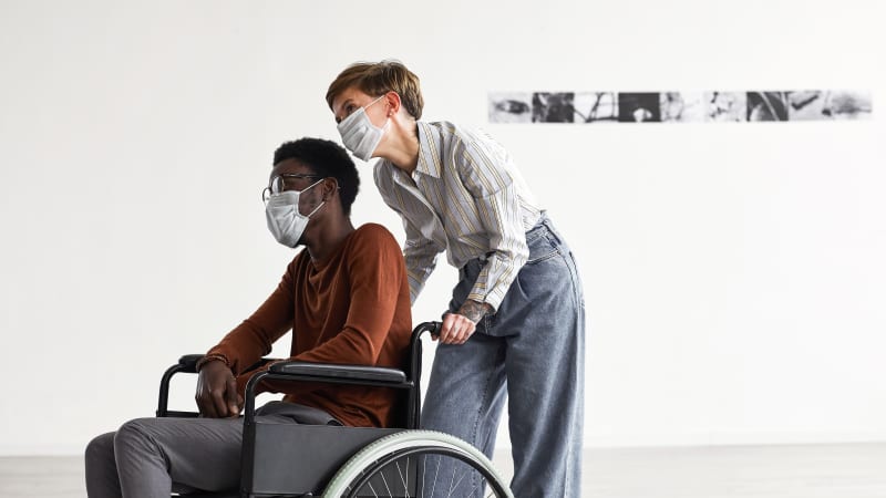 Eine junge Frau schiebt einen jungen im Rollstuhl durch eine Kunstausstellung, beide tragen medizinische Masken