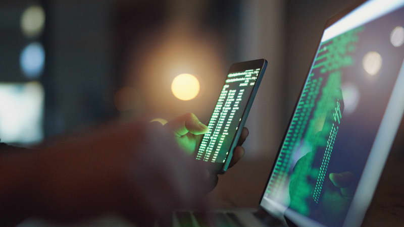 Hacker im Dunkeln mit Smartphone vor Laptop