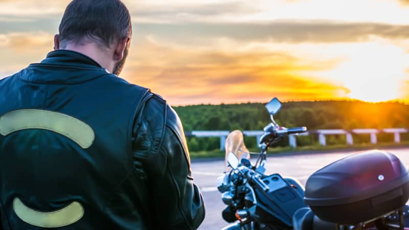 Mann in einer Lederjacke steht vor seinem Motorrad im Sonnenuntergang