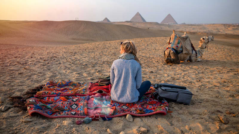 Frau sitzt auf einer Decke in der Wüste, neben ihr ein Kamel und im Hintergrund die Pyramiden von Gizeh