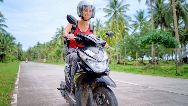 Eine junge Frau fährt mit einem Roller auf einer Straße in Thailand zwischen Palmen