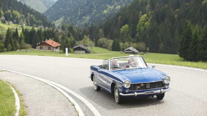 Ein älteres Paar fährt in einem blauen Fiat 1500 eine Alpenstrasse im Sommer entlang