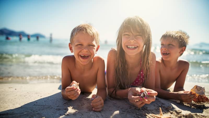 Drei Kinder liegen lachend am Strand im Wasser.