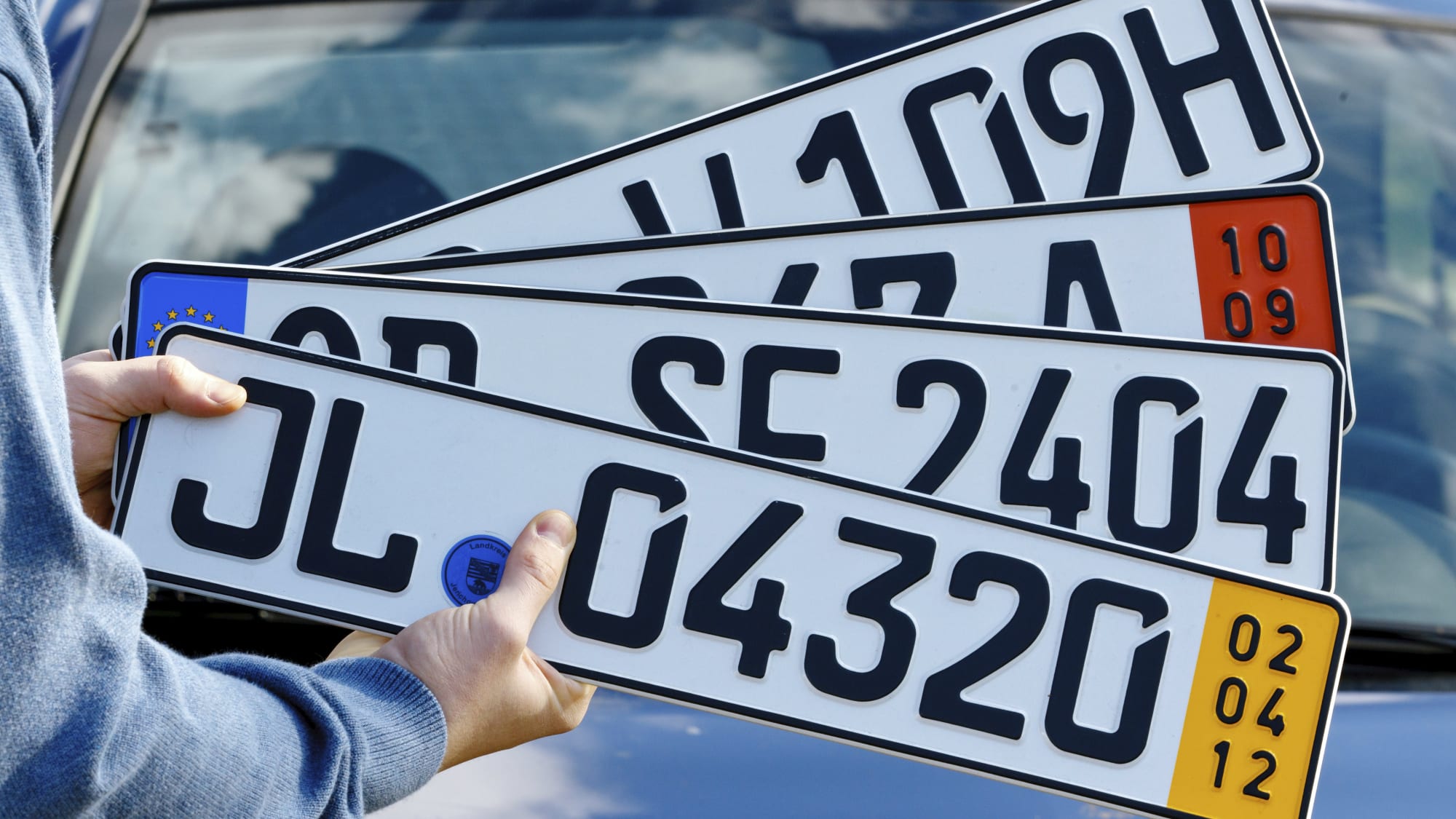 Ein Mann hält ein H-Kennzeichen für historische Fahrzeuge (v. oben), ein Ausfuhrkennzeichen, ein Euro-Kennzeichen und ein Kurzzeitkennzeichen vor ein Auto.