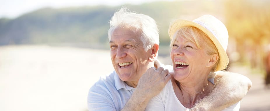 Älteres Paar schauen lachend und sich umarmend auf eine Bucht im Hintergrund: Auslandskrankenversicherung für Senioren