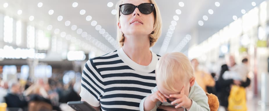 Mutter mit Kleinkind auf dem Arm steht am Flughafen und schaut auf Anzeigentafel
