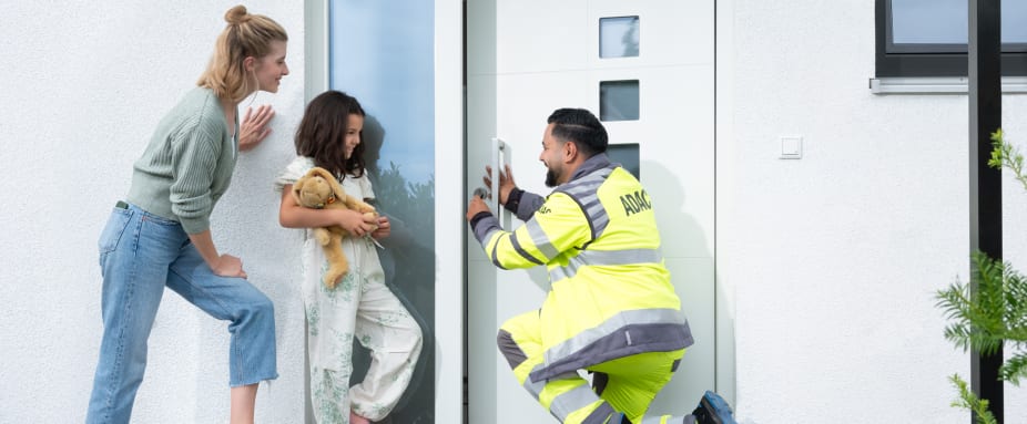 ADAC Schlüsselnotdiensthelfer öffnet kniend eine Haustür für eine ausgesperrte Mutter mit Tochter.