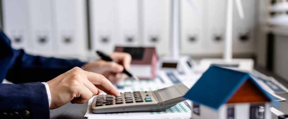 Tisch mit vielen Unterlagen, einem Modellhäuschen und einem Taschenrechner, auf dem jemand tippt