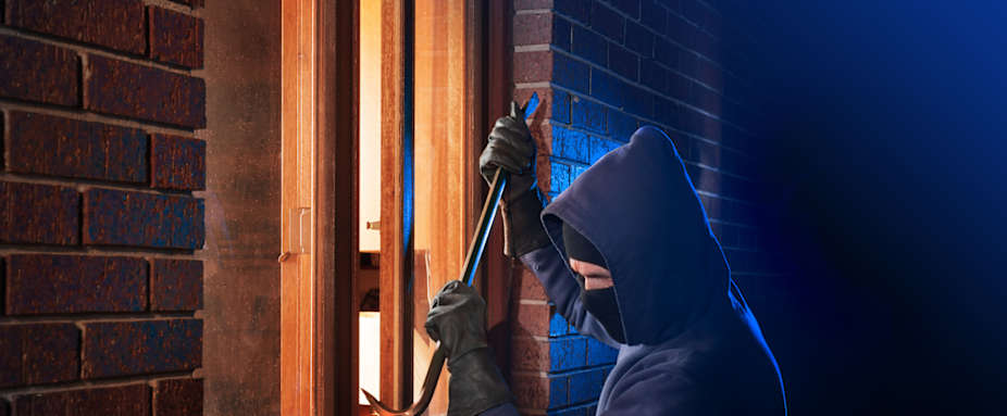 Mann in blauem Kapuzenpulli und schwarzer Skimaske bricht Fenster mit Brecheisen auf.