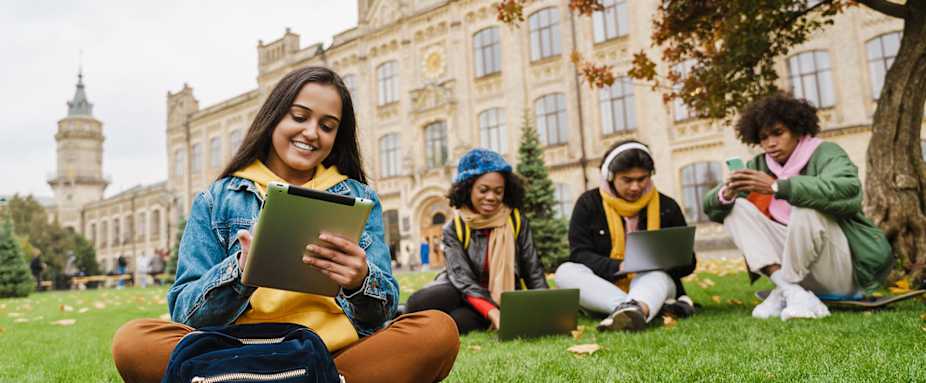 Vier Studenten sitzen vor einer alten Uni und schauen auf ihre Laptops
