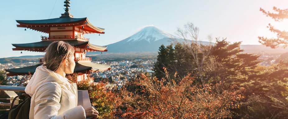 Junge Frau steht in Japan auf einer Aussichtsplatform und schaut weg von der Kamera auf einen schneebedeckten Berg, neben ihr ein Tempel