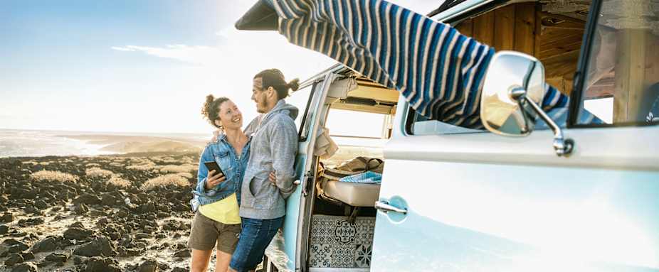 Auslandskrankenversicherung: Paar steht angelehnt am Bulli am Strand und lacht sich an