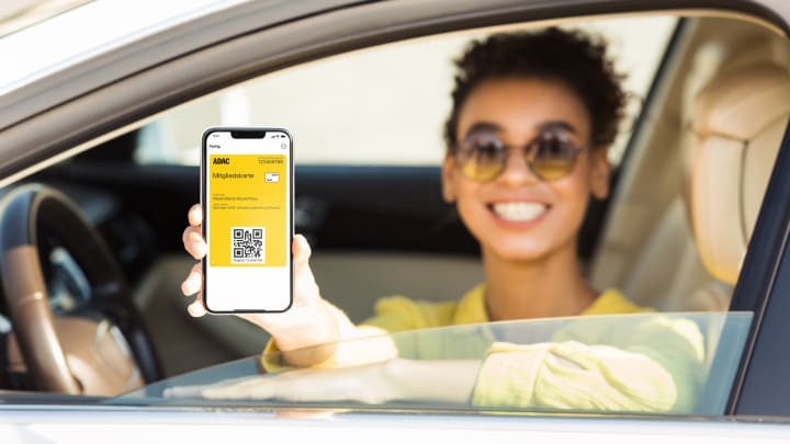 Frau zeigt aus dem Fenster ihres stehenden Autos die ADAC Mitgliedskarte auf ihrem Smartphone - ADAC Mitgliedskarte